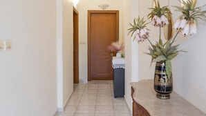A hallway with a wooden door at the end, a small table with a vase of flowers, and a wall-mounted mirror. - Lion's Yard (Chania)