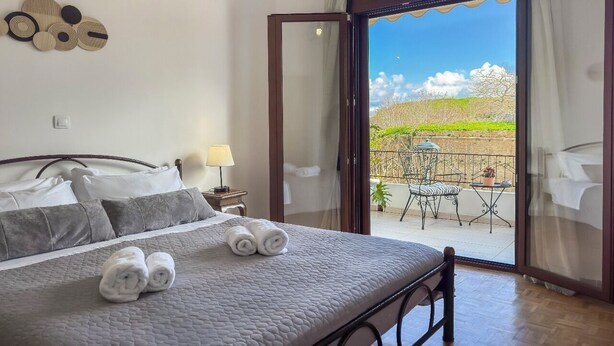 A bedroom with a bed, pillows, a bedside table with a lamp, and a view of an outdoor balcony with a table and chairs. - Lion's Yard (Chania)