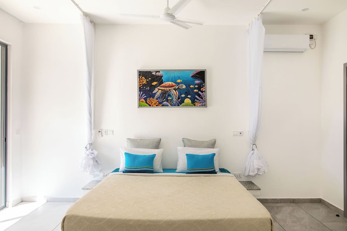 Une chambre à coucher avec un lit, deux oreillers, un ventilateur de plafond, un appareil de climatisation et un tableau représentant une tortue accroché au mur.