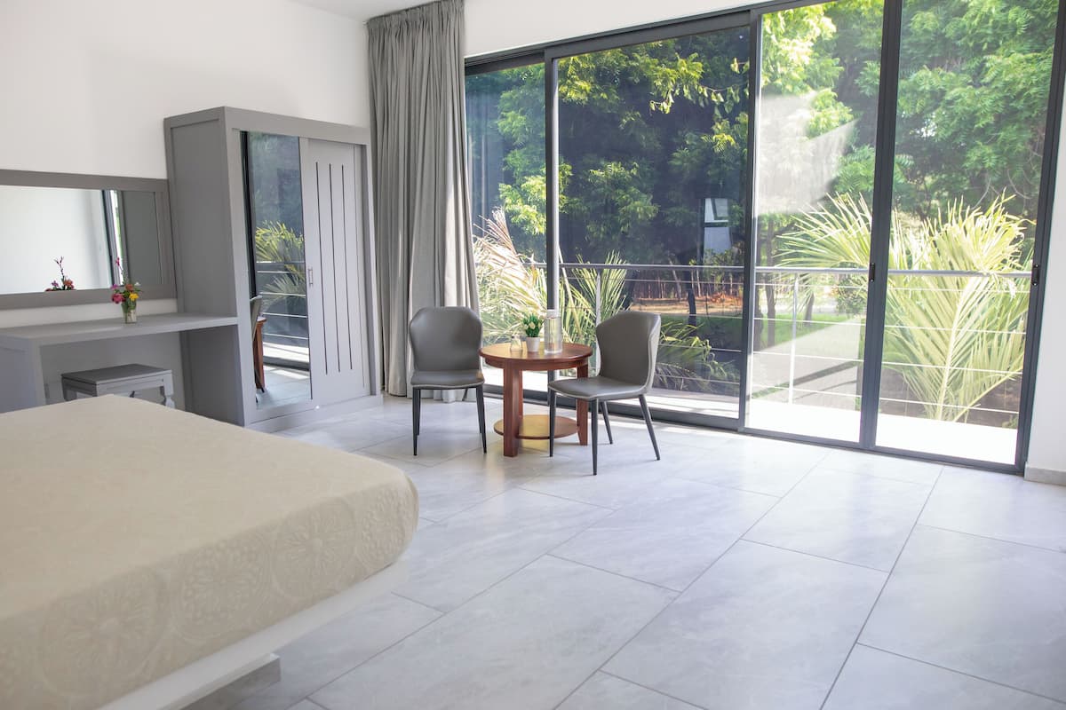 Une chambre d’hôtel moderne dotée d’un grand lit, d’un bureau, de deux chaises et d’un balcon offrant une vue sur la verdure.