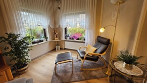 A cozy corner with a black leather chair, a small round table with a plant, and a large window with curtains. - Vacation apartment Ernebibbel - comfortable apartment with terrace (Altendiez)