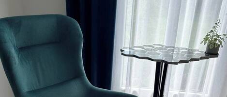 A teal armchair with a glass table beside it, a potted plant on the table, and a window with sheer curtains.