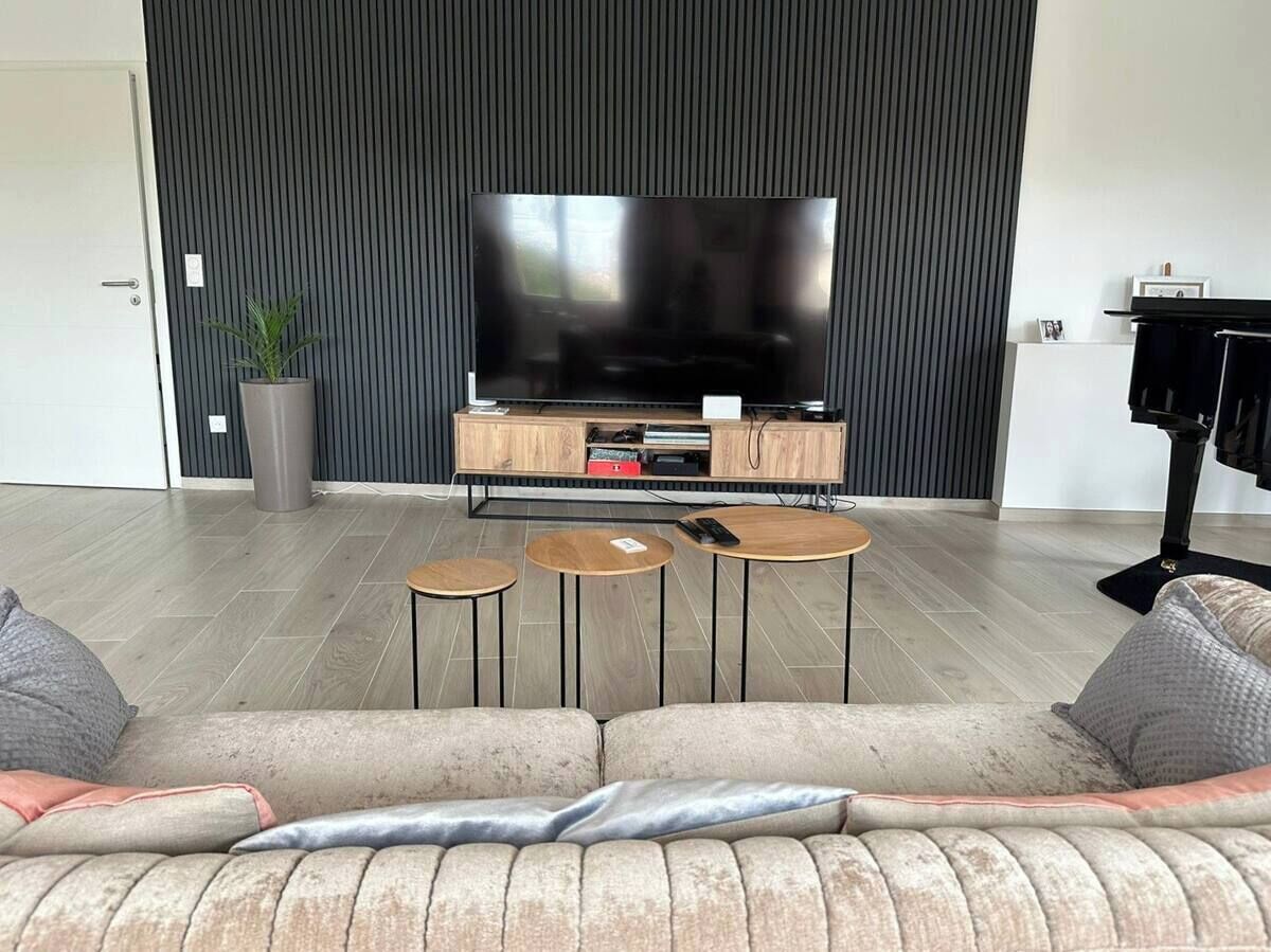 A modern living room with a large flat-screen TV, a wooden entertainment unit, and a beige sofa with cushions. There are three small round tables and a potted plant.