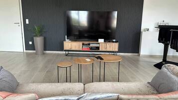 A modern living room with a large flat-screen TV, a wooden entertainment unit, and a beige sofa with cushions. There are three small round tables and a potted plant.
