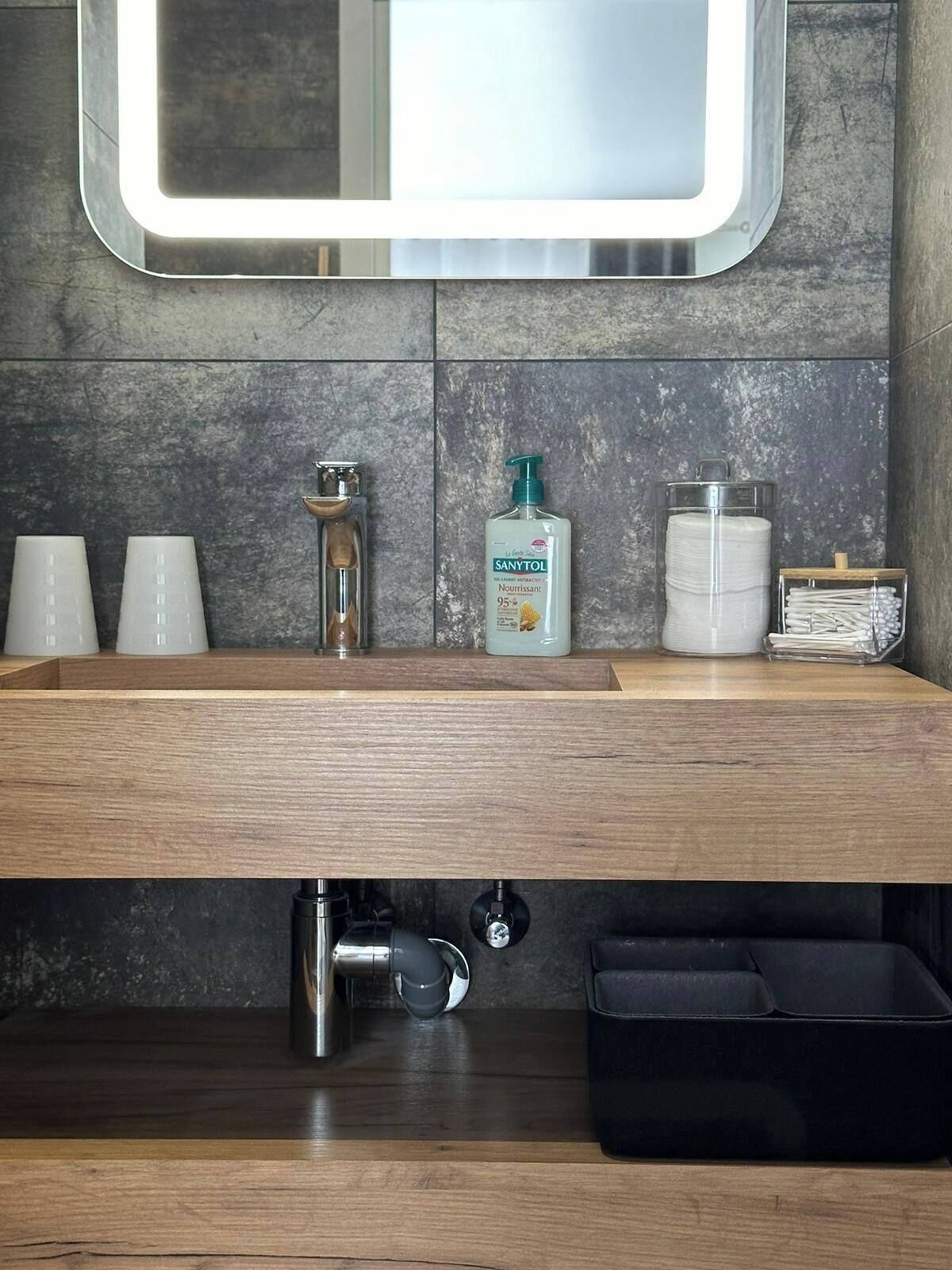 A modern bathroom with a wooden vanity, a mirror, and a sink. There are three cups on the counter, a soap dispenser, and a container of cotton swabs.