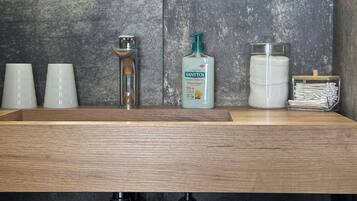 A modern bathroom with a wooden vanity, a mirror, and a sink. There are three cups on the counter, a soap dispenser, and a container of cotton swabs.
