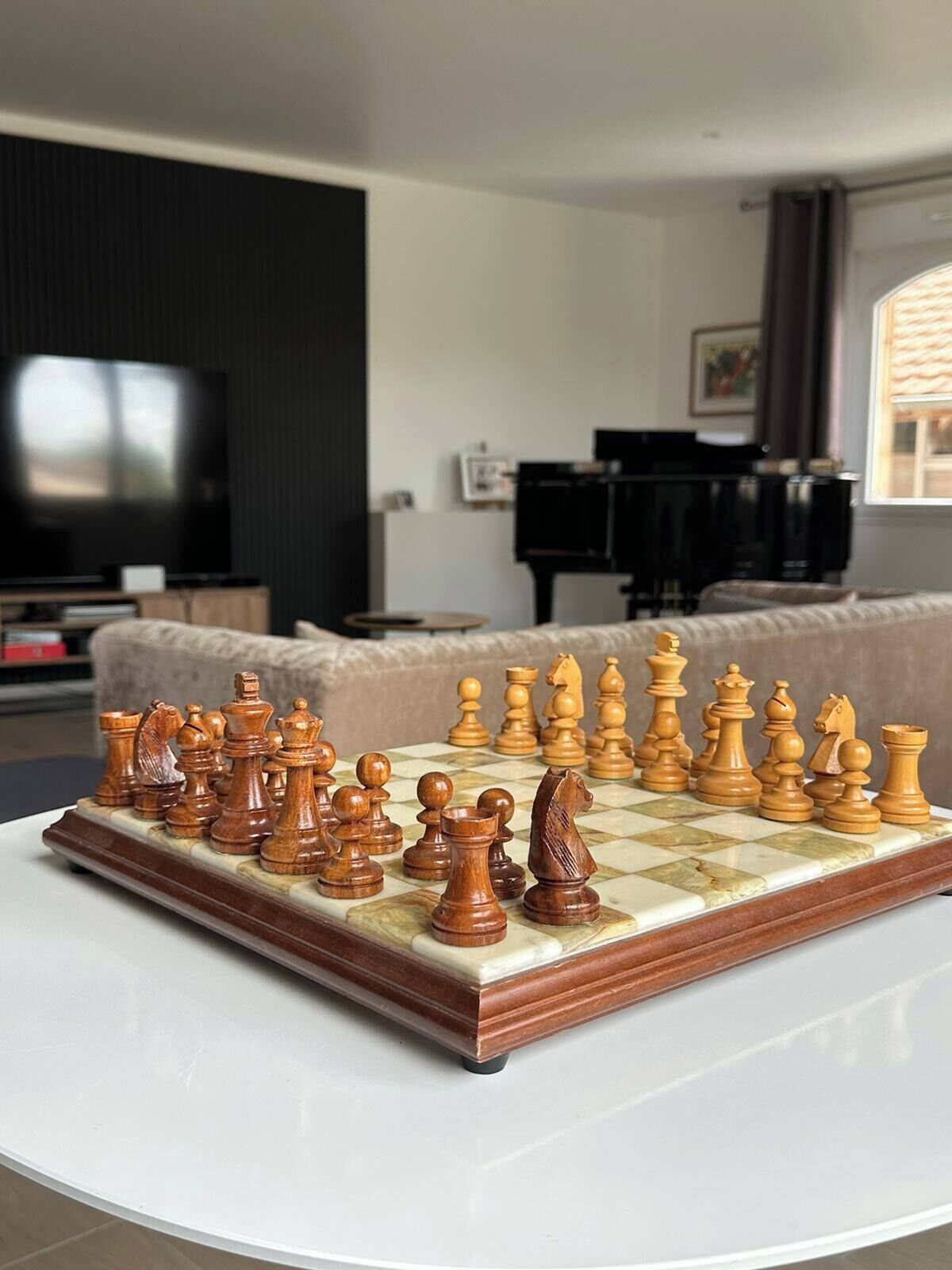 Een schaakbord op een witte tafel in een woonkamer, met een bank, een piano en een televisie op de achtergrond.