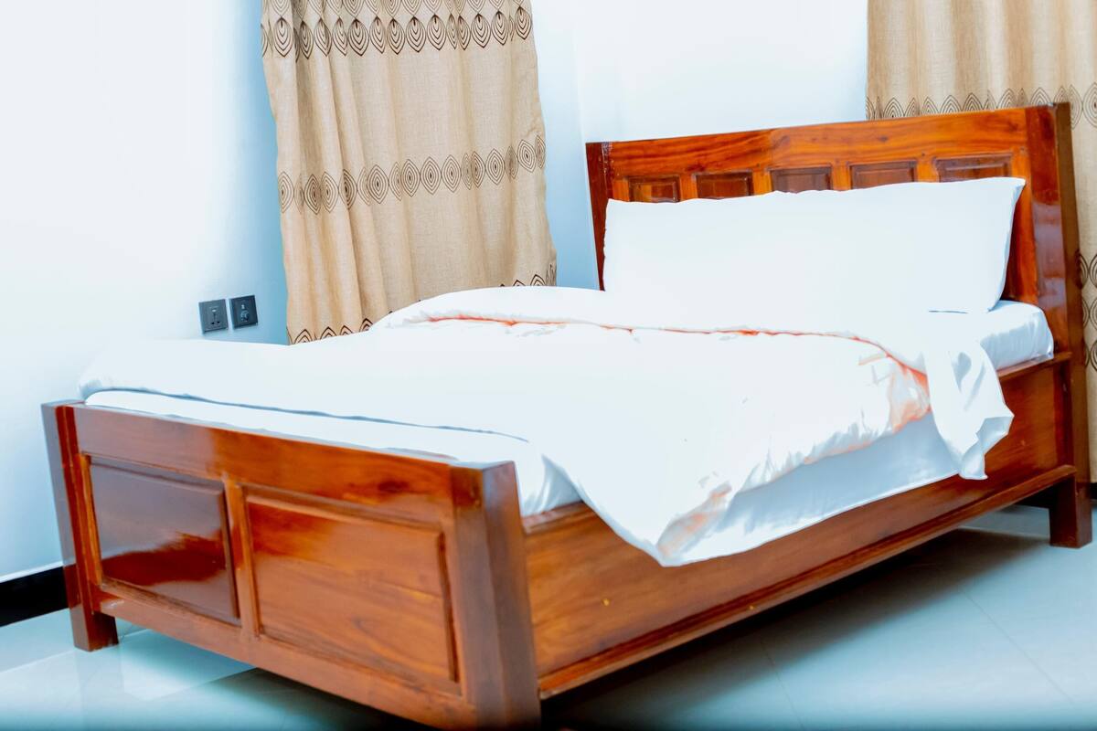 A wooden bed with a white mattress and pillows, a wooden headboard, and beige curtains with a geometric pattern.