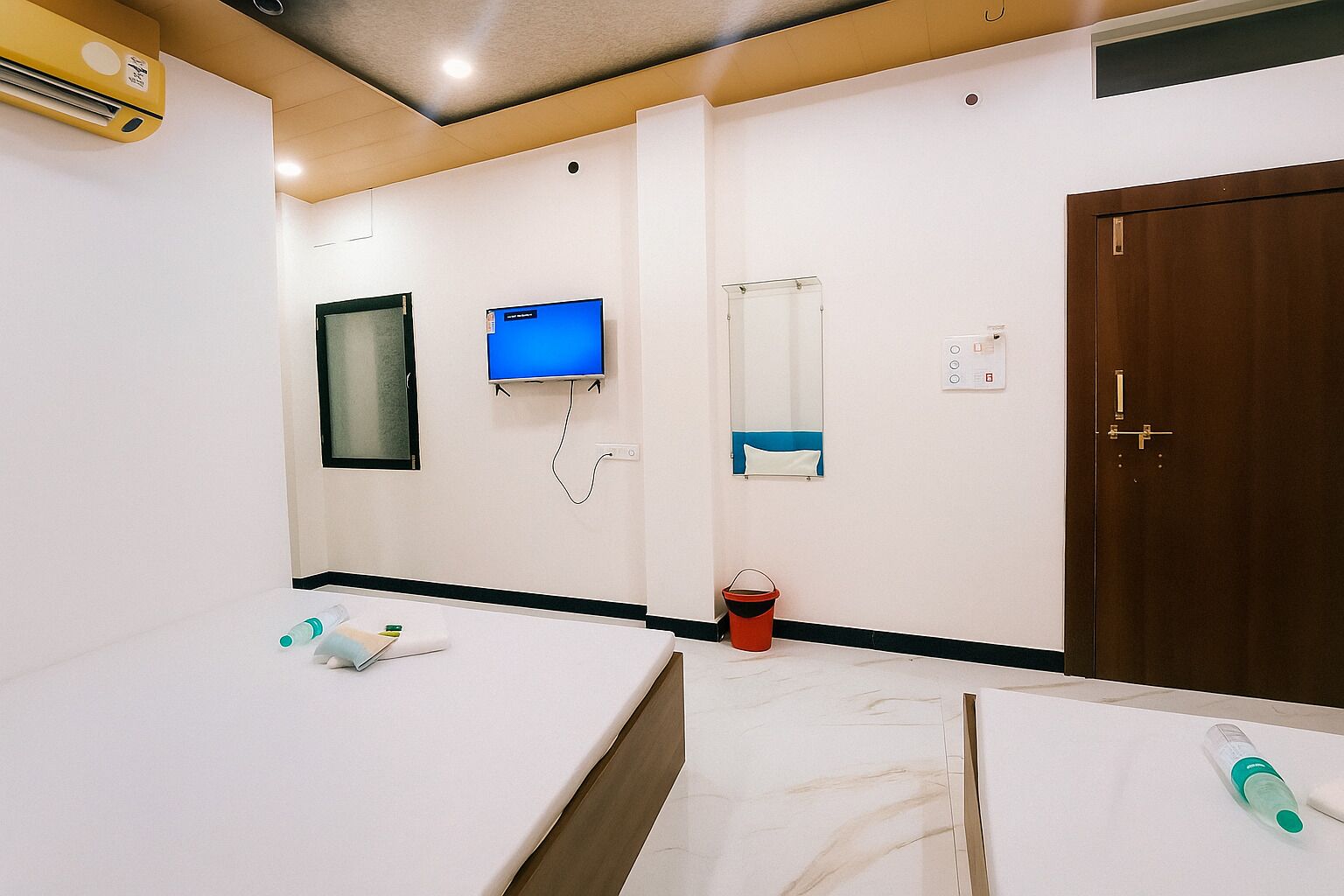 A room with a bed, a desk, a TV, a mirror, a door, and a bucket.