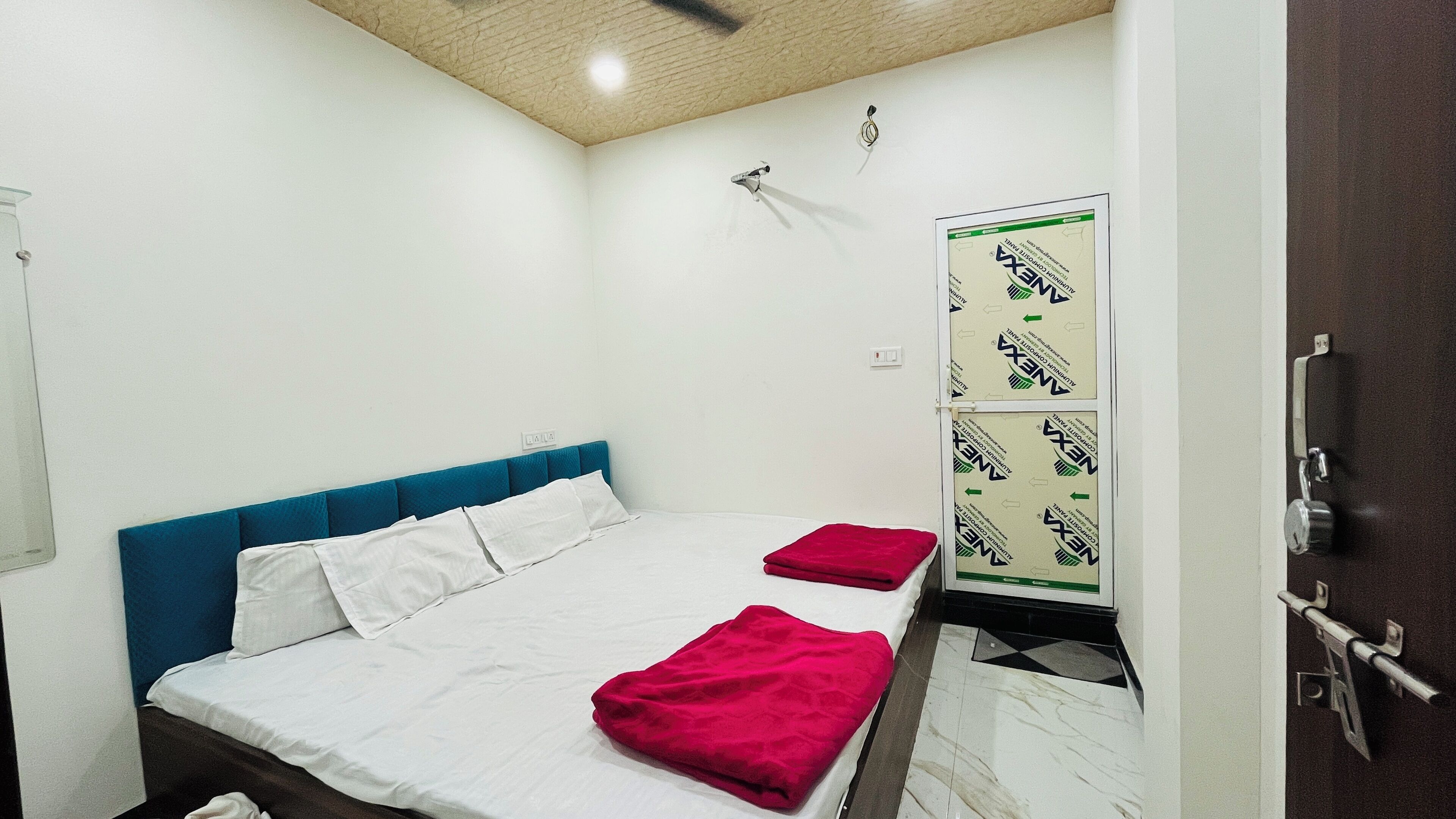 Une chambre à coucher avec un lit, une tête de lit bleue, du linge de lit blanc, une couverture rouge, un ventilateur de plafond et une porte avec une serrure.