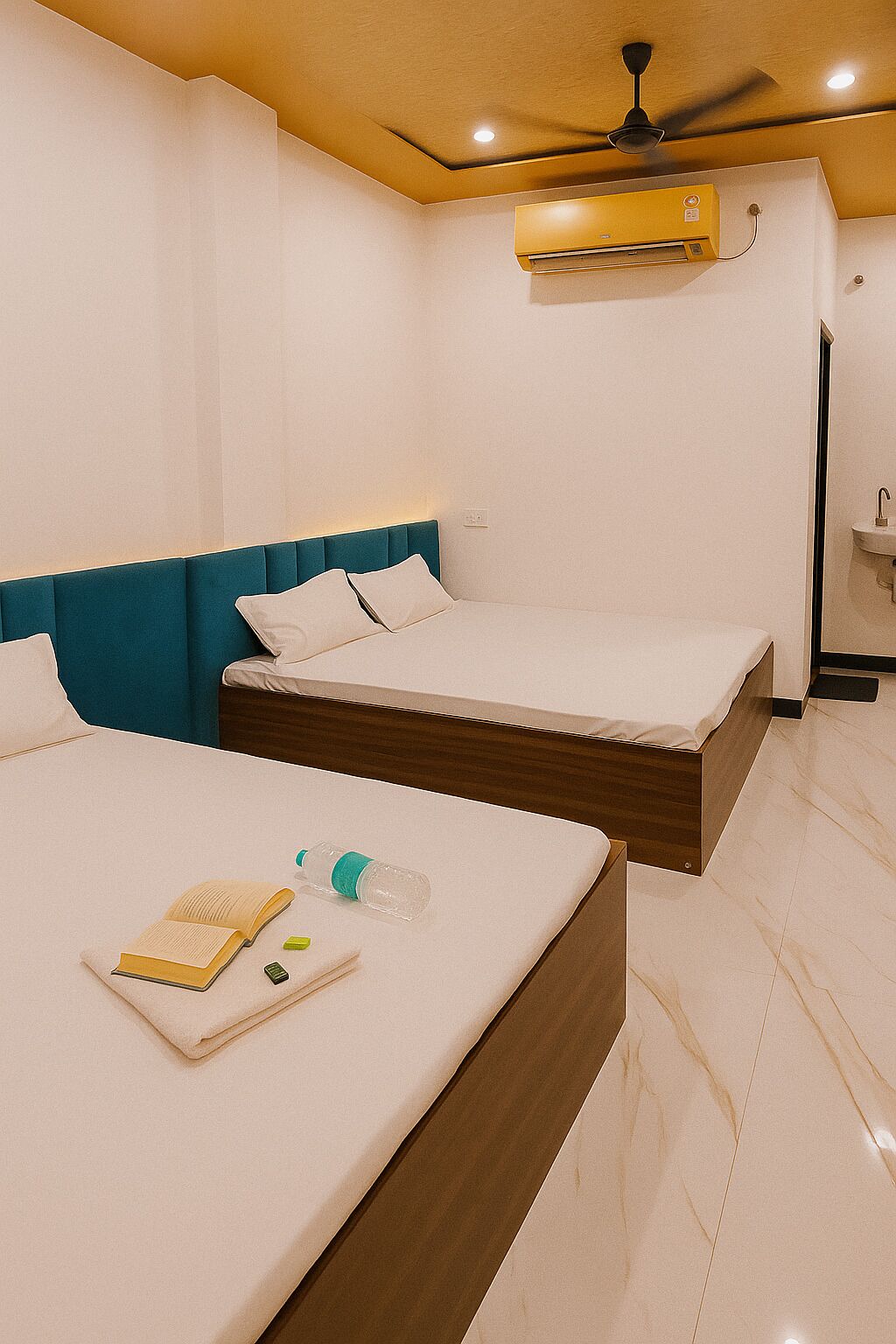 A hotel room with two beds, a ceiling fan, an air conditioning unit, and a small bathroom with a sink.