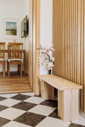 Eine hölzerne Bank mit einer Vase voller weißer Blumen in einem Raum mit einem Schachbrettmuster, Holzstühlen und einem an der Wand befestigten Fernseher.