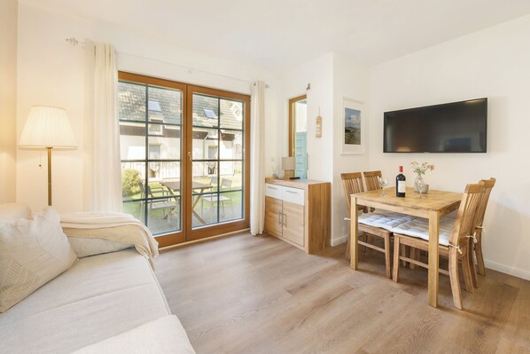 Una habitación con sofá, una mesa de comedor con sillas, televisor montado en la pared y puerta corrediza de vidrio que da a un área exterior.