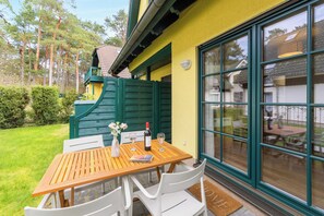 A wooden table with chairs, a bottle of wine, and a book on a patio with a yellow wall and large windows.