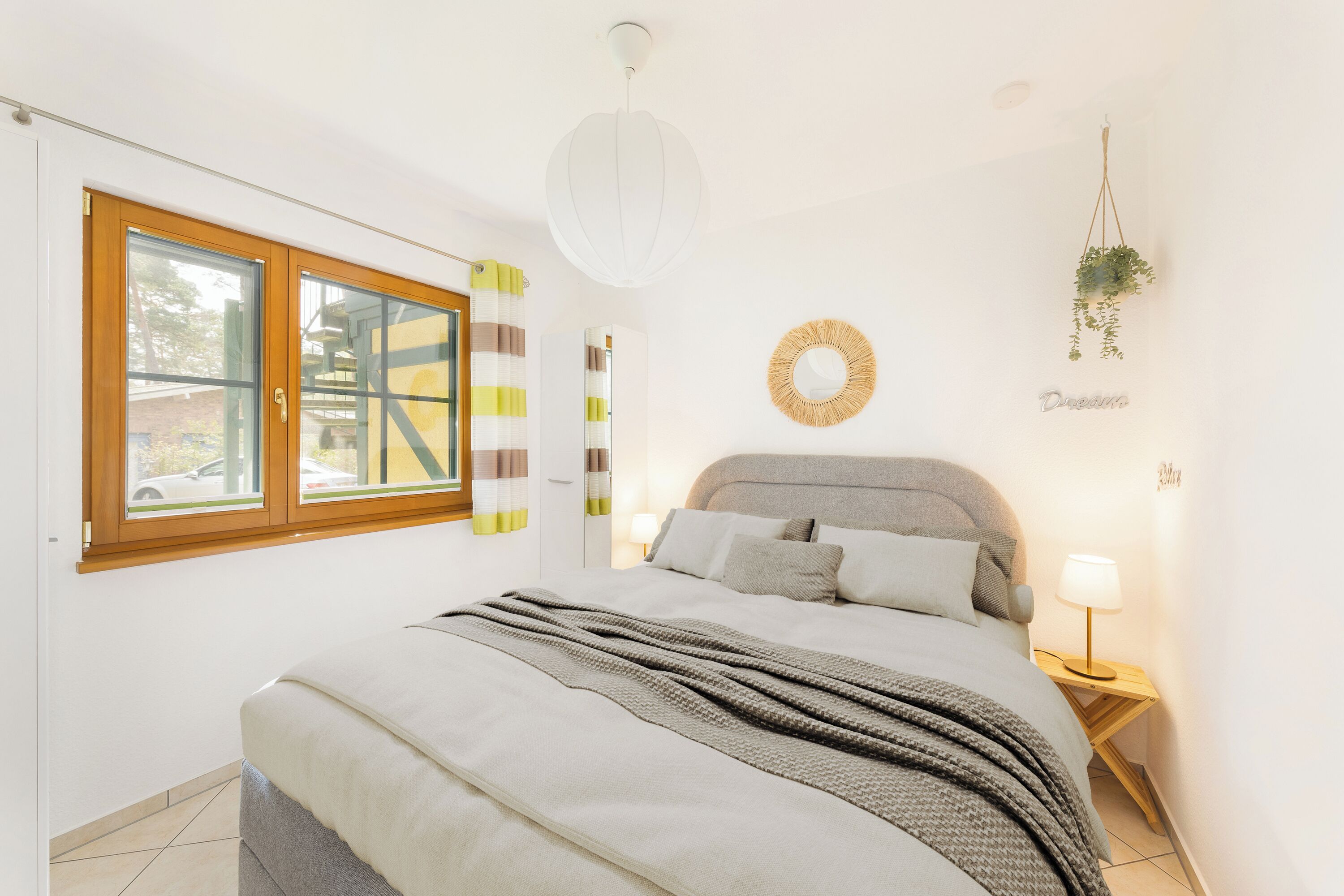 Ein Schlafzimmer mit einem Bett, einem Nachttisch mit Lampe, einer hängenden Pflanze, einem Wandspiegel und einem Fenster mit Vorhängen.