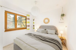 Ein Schlafzimmer mit einem Bett, einem Nachttisch mit Lampe, einer hängenden Pflanze, einem Wandspiegel und einem Fenster mit Vorhängen.