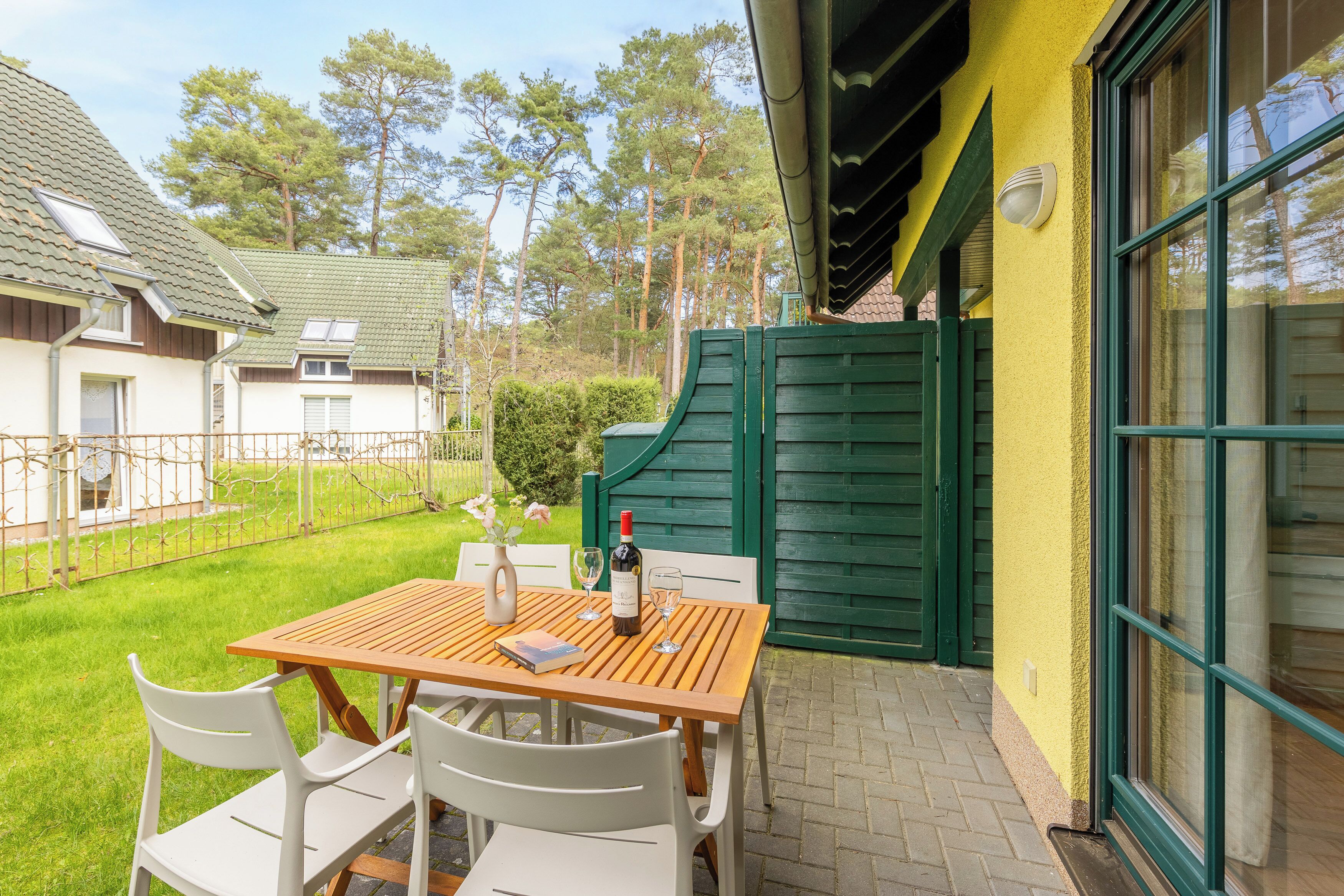 Ein Holztisch mit Stühlen und einer Weinflasche, auf einer Terrasse mit einem grünen Tor und einer gelben Wand im Hintergrund.