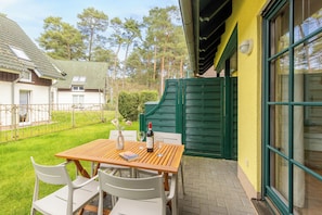 A wooden table with chairs and a bottle of wine, set on a patio with a green gate and a yellow wall in the background.