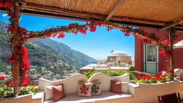 A balcony with a white cushioned seating area, a view of a mountainous landscape, and a building with a dome structure in the background. - YourHome - La Casupola (Positano)