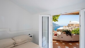 A bedroom with a bed, a small bedside table, and a sliding glass door leading to a balcony with a view of a coastal town and mountains. - YourHome - La Casupola (Positano)