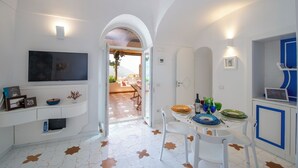 A modern, well-lit room with a TV, a dining area with a table set for two, and a view through an open door to an outdoor seating area. - YourHome - La Casupola (Positano)