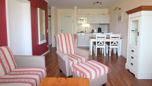 Un salon comprenant un canapé, un fauteuil et un pouf, un coin repas avec une table et des chaises, et une cuisine avec des meubles blancs et des appareils électroménagers.