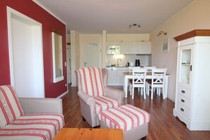 A living room with a sofa, armchair, and ottoman, a dining area with a table and chairs, and a kitchen with white cabinets and appliances. - The spacious Comfort apartment offers pure relaxation. (Ahrenshoop)