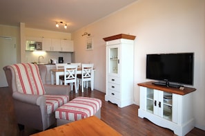 Ein kompakter Wohnbereich mit Sofa, Hocker, Esstisch, Stühlen, einer Schrank mit Glastüren, einem Fernseher und einer Küchenzeile mit Schränken und einer Mikrowelle.