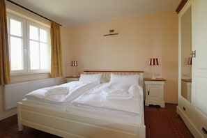A hotel room with a double bed, two bedside tables with lamps, a radiator, and a window with curtains. - The spacious Comfort apartment offers pure relaxation. (Ahrenshoop)