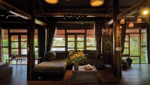 A cozy room with a sofa, a wooden coffee table, and a vase of flowers. There are large windows with curtains, and a wooden ceiling with hanging lights. - Mat Retreat (Hanoi)