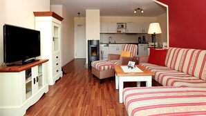 A living room with a television, sofa, ottoman, and a small table. There is a kitchen in the background with cabinets and a stove. - The particularly spacious Apartment Suite offers pure relaxation. (Ahrenshoop)