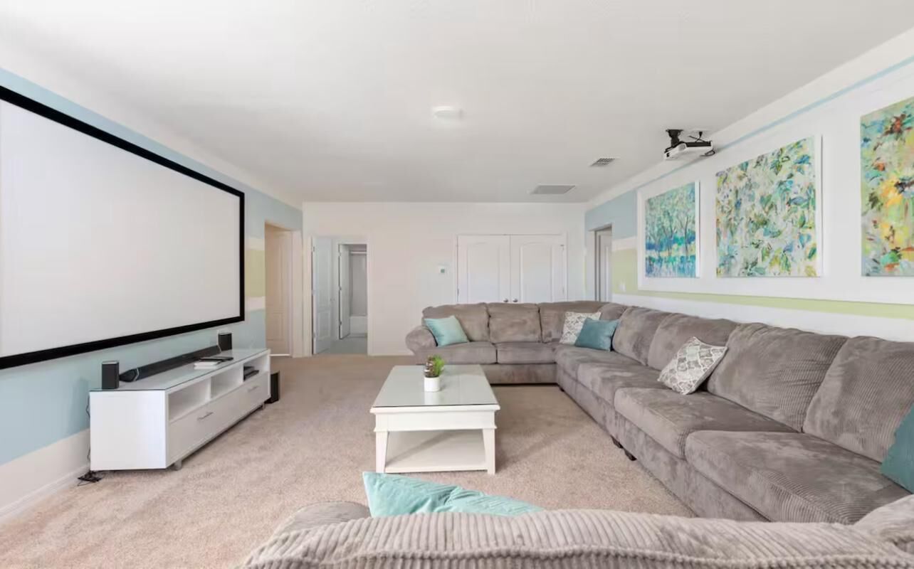 A living room with a large flat-screen TV, a gray sectional sofa, a white coffee table, and a wall-mounted projector.