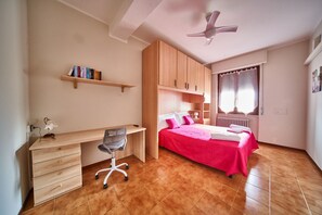Une chambre avec un bureau en bois, une chaise, un lit recouvert d’une couverture rose, un ventilateur de plafond et une fenêtre avec des rideaux.