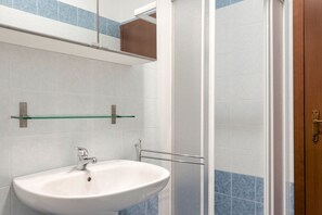 Ein Badezimmer mit weißem Waschbecken, einer Glasablage darüber und einer Duschkabine mit blauen Fliesen.