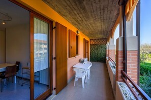 A covered outdoor area with a table and chairs, a wooden door, and a view of greenery.
