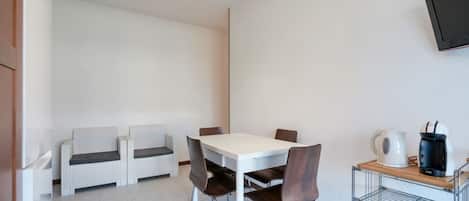 A small, modern room with a white table and four chairs, a sofa, a TV mounted on the wall, and a kettle on a shelf.