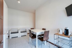 A small, modern room with a white table and four chairs, a sofa, a TV mounted on the wall, and a kettle on a shelf.