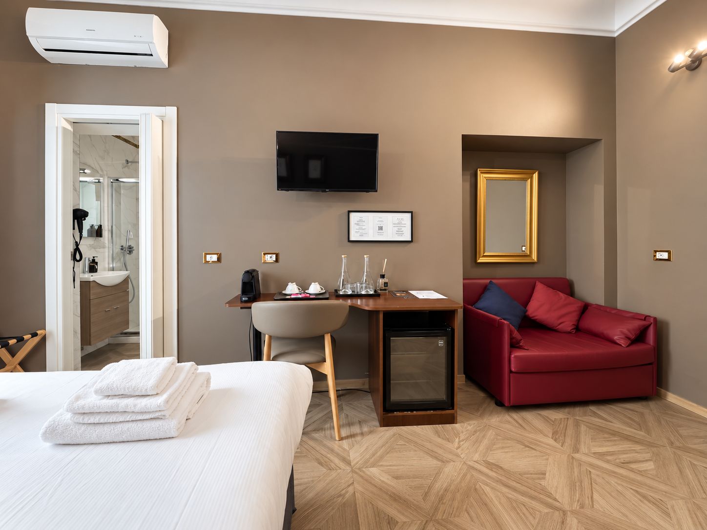 A hotel room with a bed, a desk with a chair, a red sofa, a television, and a mirror.