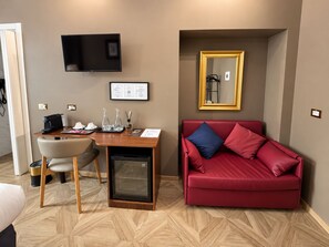 A compact room with a wooden desk, a red sofa, a television, and a small refrigerator. - Whim Collection Vaticano Prati  (Rome)