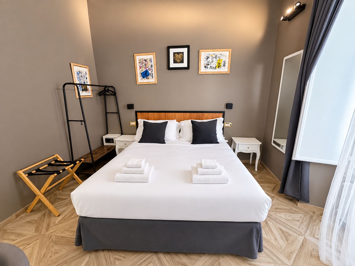 Een hotelkamer met een groot bed, twee nachtkastjes, een wandplank met kunstwerk, een houten vloer en een raam met gordijnen.