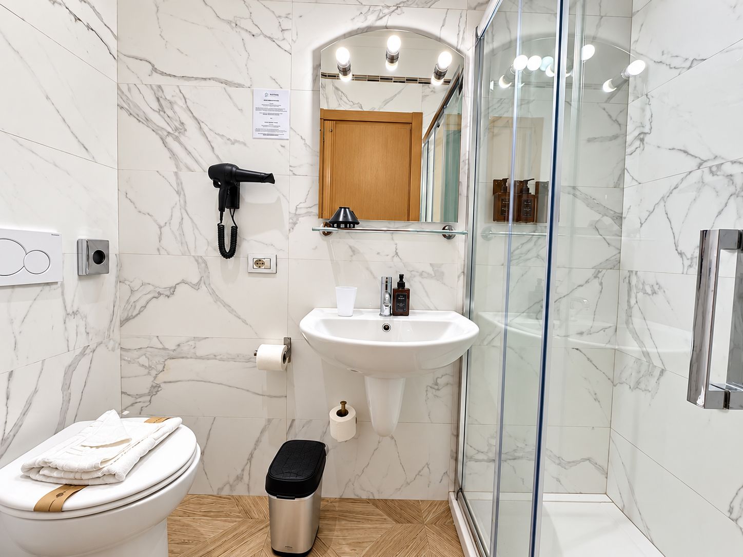 A modern bathroom with a marble floor, a white sink, a glass shower enclosure, a toilet, and a hairdryer.