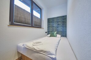 Una cama pequeña y bien arreglada con sábanas blancas y dos toallas, una ventana con persianas, y un cabecero azul y blanco con diseño.