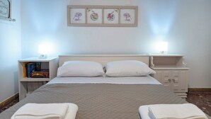 Un lit bien fait, recouvert d’une courtepointe, deux tables de chevet avec des lampes, une œuvre d’art florale encadrée au-dessus du lit et une bibliothèque remplie de livres.