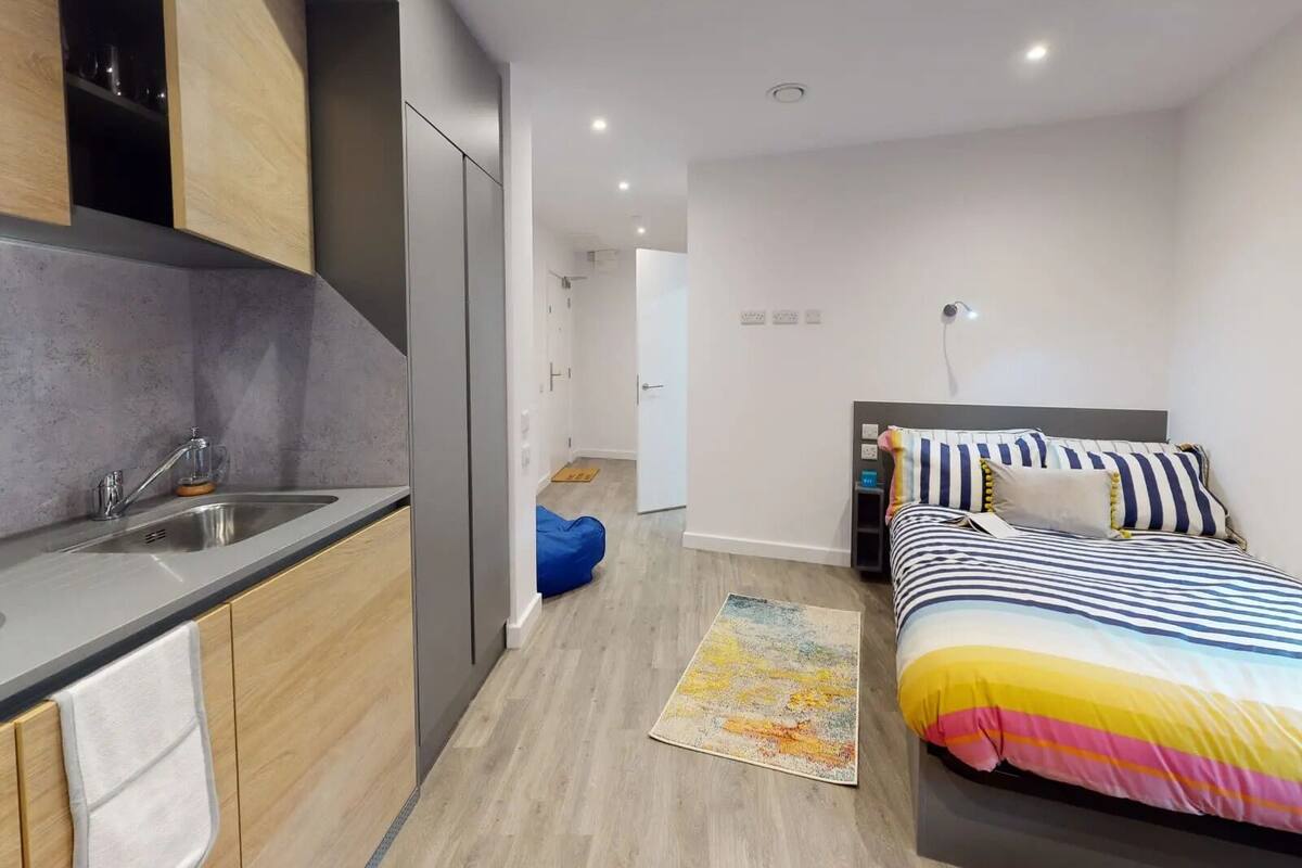 A modern hotel room with a kitchen area, a bed with striped bedding, and a colorful rug.