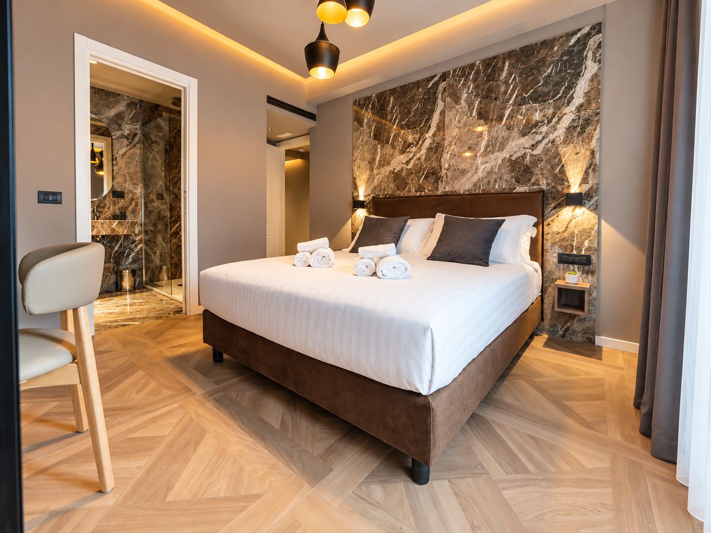 Quarto de hotel com uma cama grande, área de estar com uma poltrona, parede de mármore e banheiro visível através de uma porta aberta.