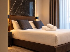 A neatly made bed with white linens and two dark pillows, a marble headboard, and a window with curtains. - Whim Collection Trastevere (Rome)