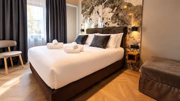 A modern hotel room with a large bed, a wooden floor, a stone accent wall, and a seating area with a chair and ottoman.