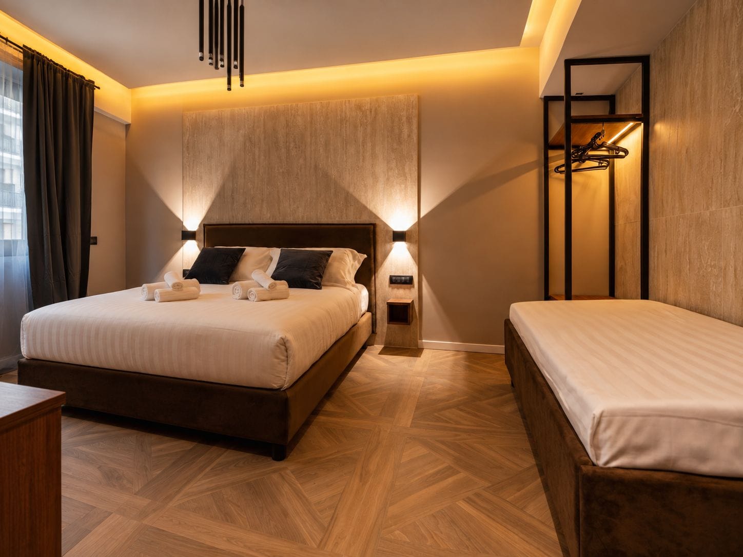 Una habitación de hotel moderna con una cama grande, una cama más pequeña, una mesita de noche, una lámpara de pared y suelo de madera.