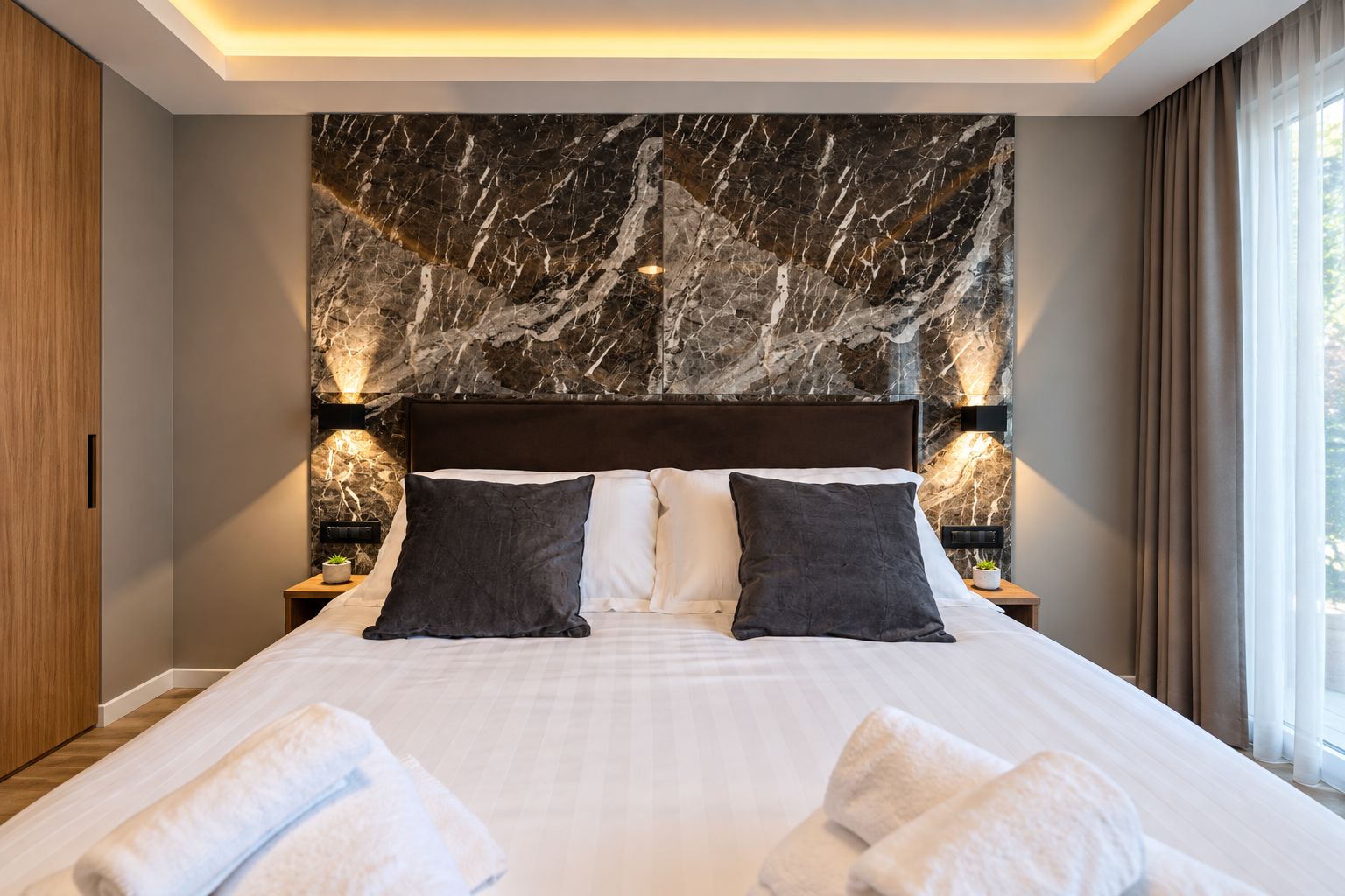 A hotel room with a large bed, marble headboard, two bedside tables with lamps, and a view of the outdoors through a window.