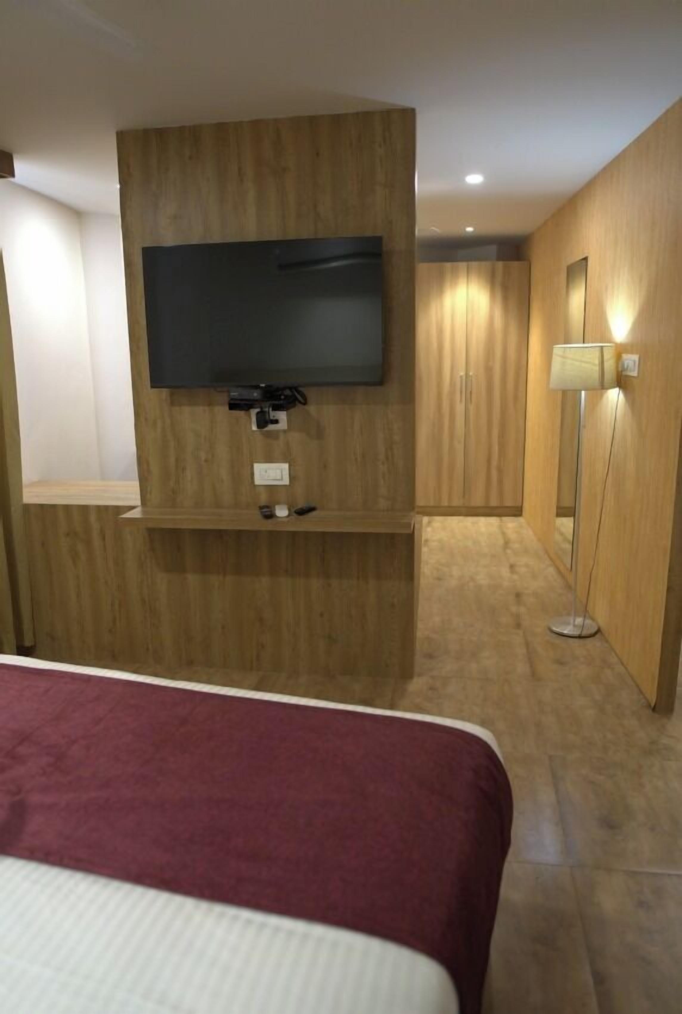 A hotel room with a bed, a mounted TV, a bedside table, a lamp, and wooden paneling on the walls.